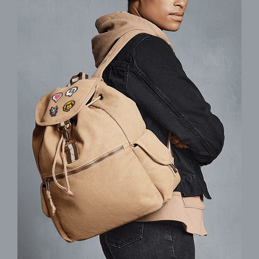 A person wears a tan canvas Stardew Valley backpack adorned with several embroidered patches including a heart emote and cat design.