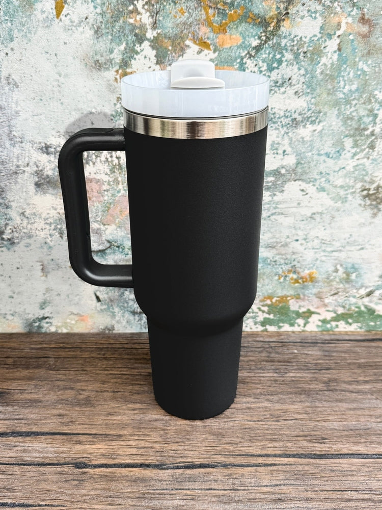 A black 40oz D&D engraved tumbler sits on a wooden surface against a textured wall.
