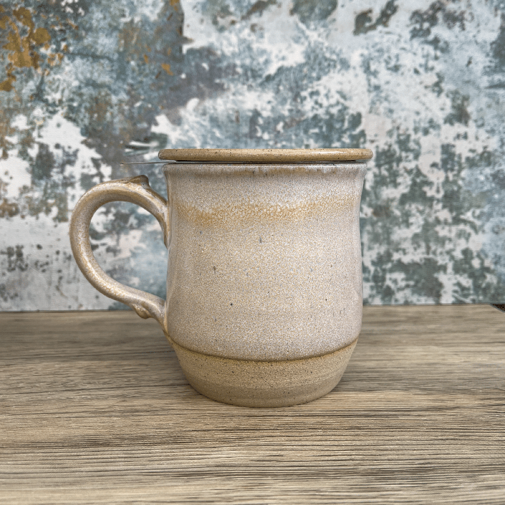 A handmade ceramic tea infuser mug with a lid sits on a wooden surface against a textured blue and white background.