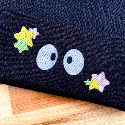 A black Soot Sprites beanie features embroidered eyes and colorful stars inspired by Ghibli's Susuwatari.
