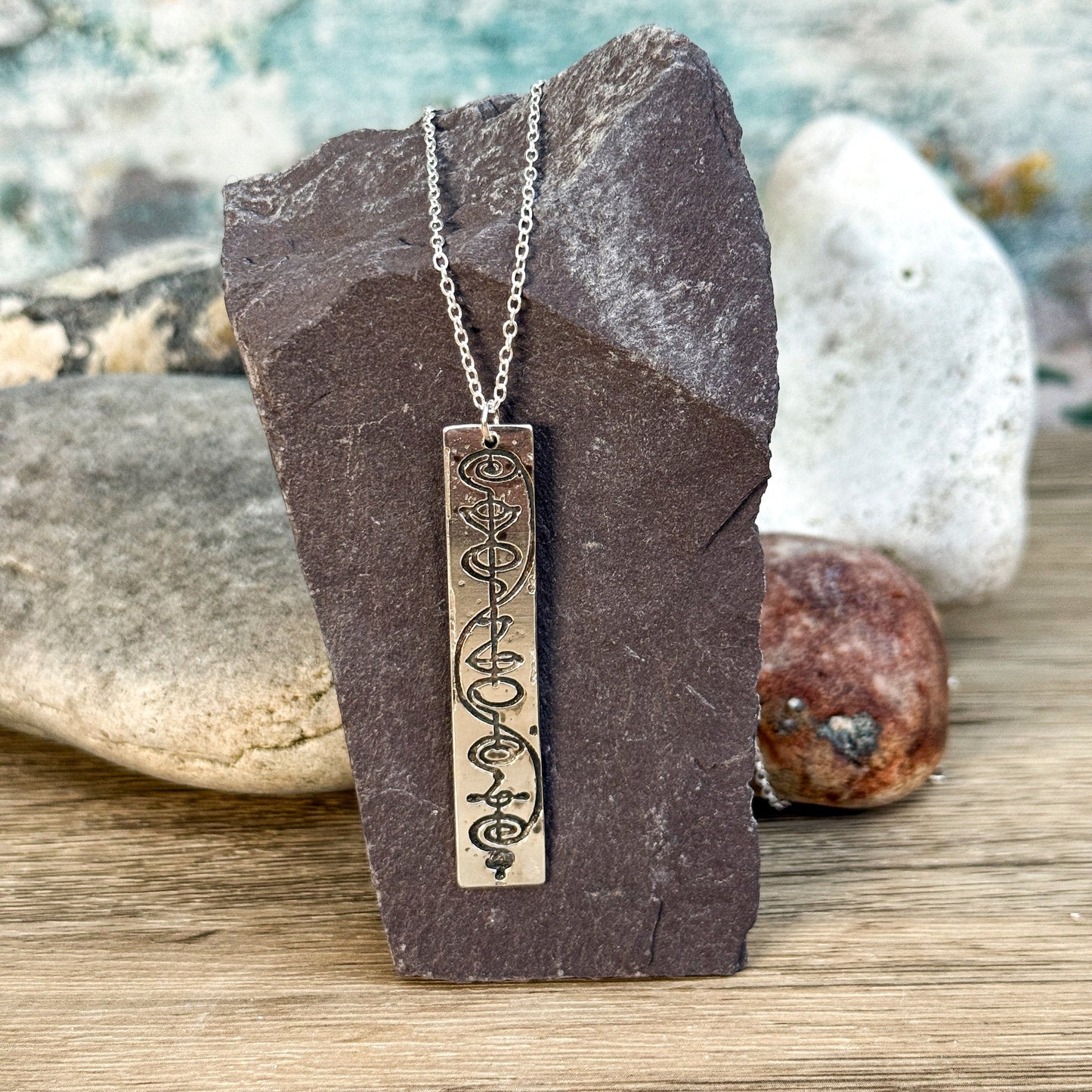 The Quirkify Star Trek Vulcan IDIC Calligraphy Sterling Silver Pendant Necklace features an intricate rectangular design, elegantly displayed on smooth stones with a softly gleaming finish against a muted background.