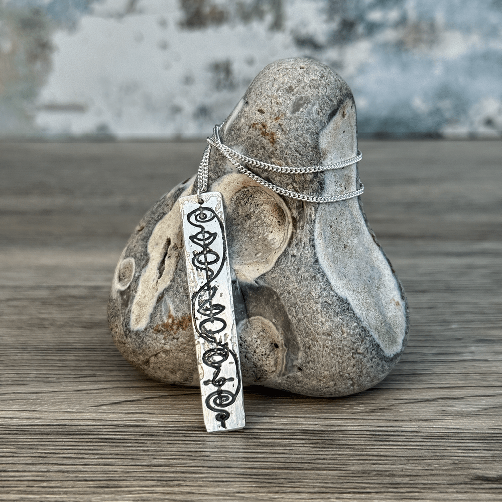 A sterling silver Vulcan IDIC pendant necklace features calligraphy etching on a rectangular charm suspended from a delicate chain against a rustic background.