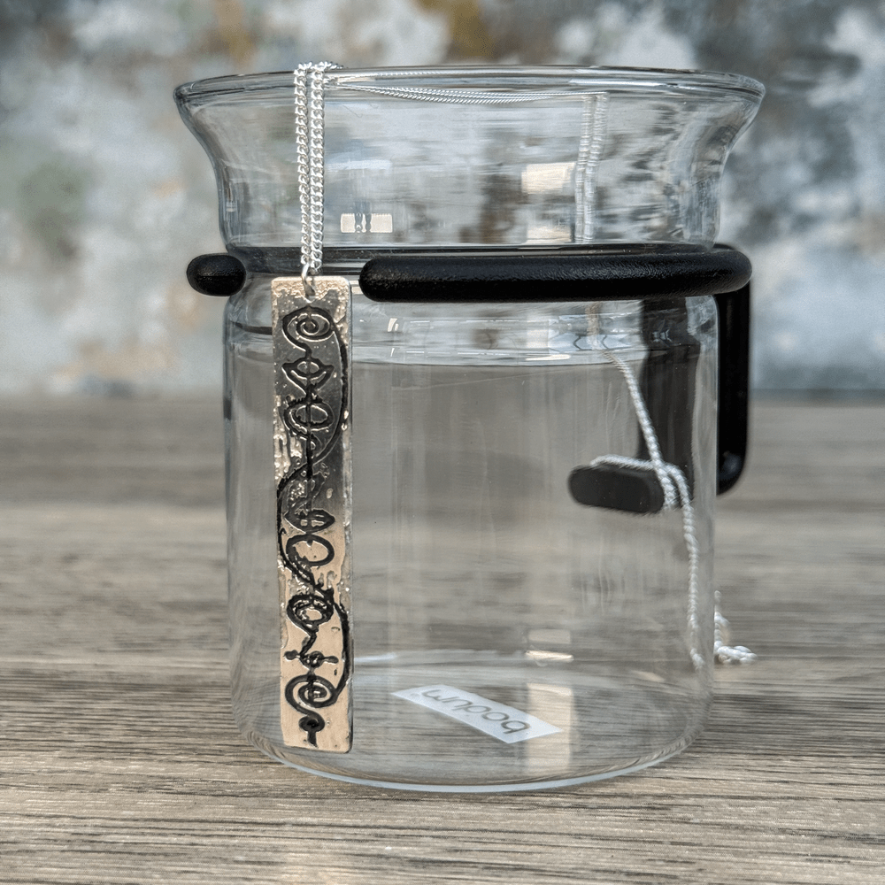 A Sterling Silver Vulcan IDIC pendant necklace hangs from a delicate chain, displayed against a glass tea infuser on a wooden surface.