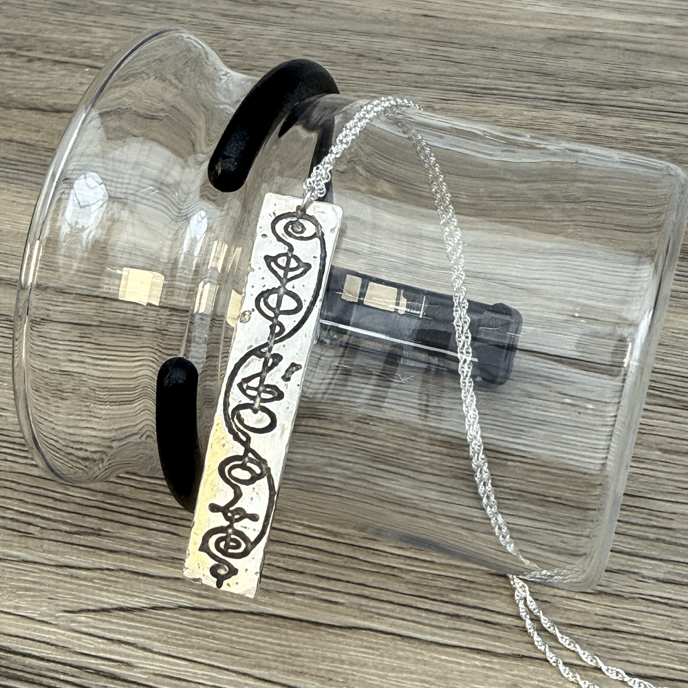 A sterling silver Vulcan IDIC calligraphy etched pendant hangs from a delicate chain, displayed against a clear glass container on a wooden surface.