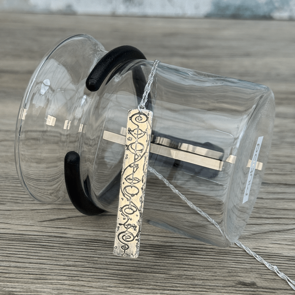 A sterling silver Vulcan IDIC pendant necklace featuring calligraphy etching is displayed draped over a glass beaker.