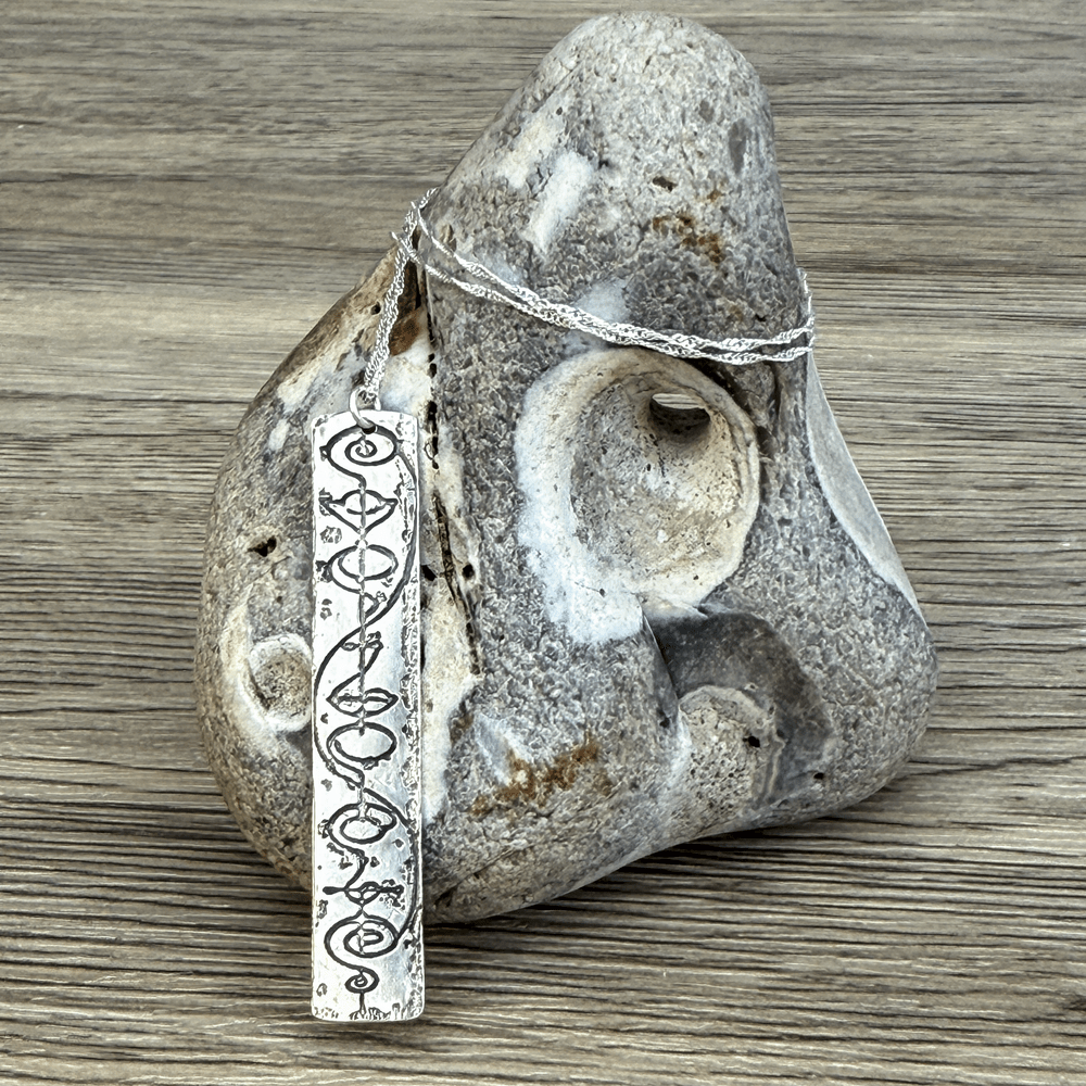 A sterling silver Vulcan IDIC pendant necklace featuring calligraphy etching hangs against a textured stone surface.