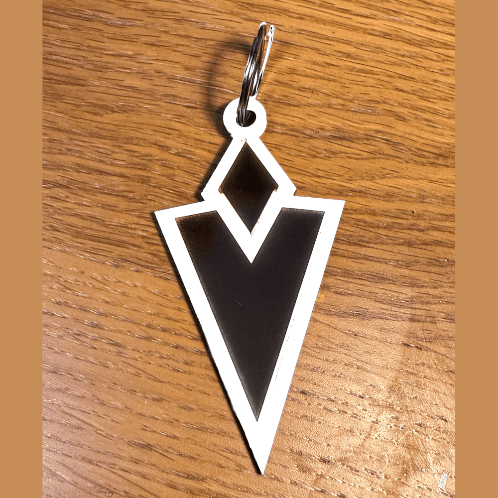 A 3D acrylic keyring featuring a black and white Skyrim inspired Quest Marker icon resting on a wooden surface.