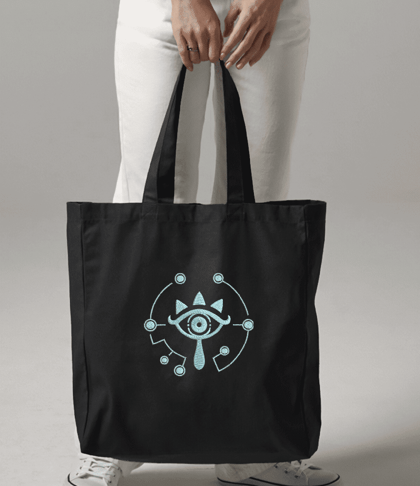 A black canvas tote bag featuring a light blue embroidered Sheikah eye symbol from Zelda.