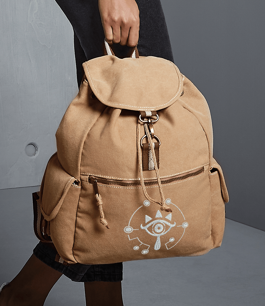 A tan Zelda Sheikah backpack features glow-in-the-dark embroidery of a circular symbol on the front pocket.