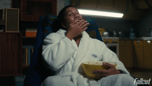 A person wearing a white bathrobe embroidered with the Fallout logo sits in an armchair eating popcorn.