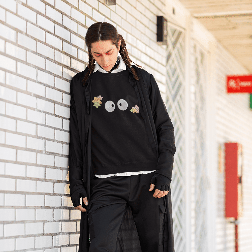 A person wearing a black Soot Sprites embroidered sweater inspired by Studio Ghibli stands against a brick wall.
