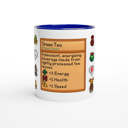 A white mug with a blue rim features a Stardew Drinks pattern depicting a Green Tea beverage card with pixelated sprites around its border.