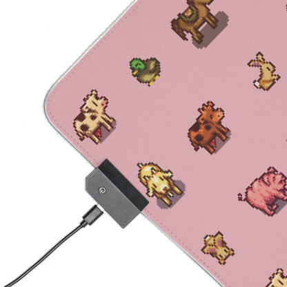 A pink desk mat features pixel art depictions of animals from Stardew Valley, including a cow, duck, and pig, and is connected to a power source via USB cable.