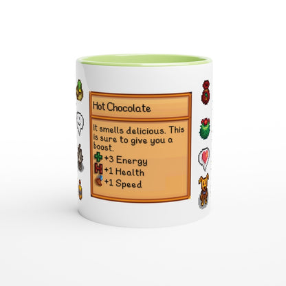 A white mug with a green rim features a Stardew Drinks pattern depicting a hot chocolate beverage card with pixelated sprites and stats.