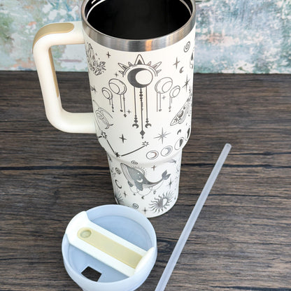 A tall, white Witchy Boho engraved tumbler featuring celestial magic designs of stars, moons, and crystals sits on a wooden surface alongside its lid and straw.