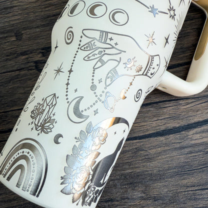 A cream-colored engraved tumbler features celestial designs including stars, moons, crystals, and a rainbow, reflecting a witchy boho aesthetic.