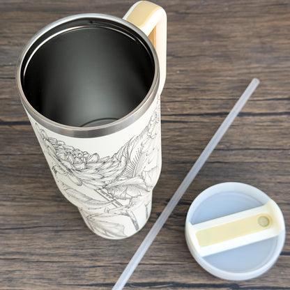 A white Floral Dahlias 40oz Tumbler features a unique engraved botanical design of dahlias and leaves on its surface, alongside a straw and lid resting on a wooden table.