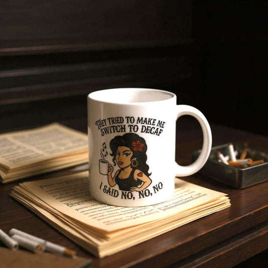 A white coffee mug featuring an illustration of a woman and text referencing "No to Decaf" is displayed on vintage books and stationery.