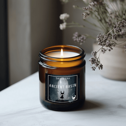A lit amber glass candle labeled "Ancient Basin" referencing the Hollow Knight video game sits on a white marble surface next to dried flowers.