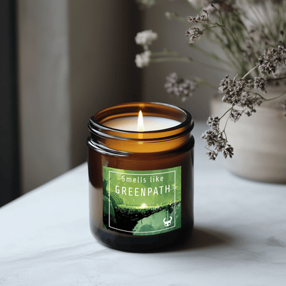 A lit candle in an amber jar displays a label that reads "Smells like GREENPATH" and features imagery inspired by the video game Hollow Knight.