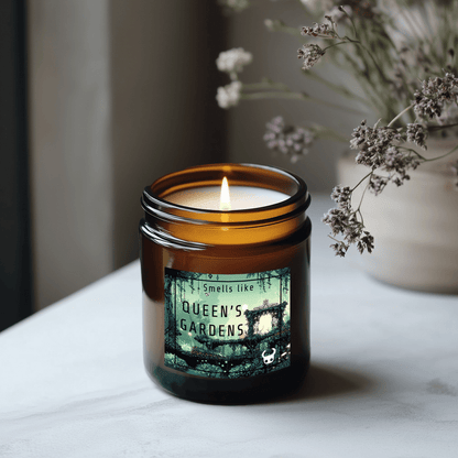 A lit candle in a brown glass jar displays a label reading "Smells like Queen's Gardens" and featuring imagery inspired by Hollow Knight.