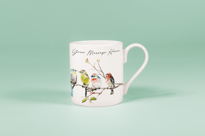 A white china mug displays watercolour birds perched on a branch alongside handwritten text reading "Your Message Here.