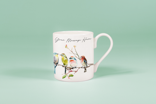 A white china mug displays watercolour birds perched on a branch alongside handwritten text reading "Your Message Here.