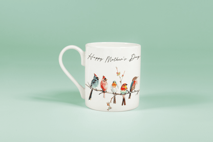 A white china mug displays watercolour birds perched on a branch alongside handwritten text reading "Happy Mother's Day".