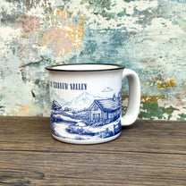 The Quirkify Stardew Valley Chinoiserie Campfire Mug is a blue and white toile ceramic cup featuring house, tree, and mountain artwork with "Stardew Valley" printed on it, displayed on a wooden surface against a multicolored wall.