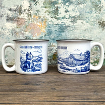 The Quirkify Stardew Valley Chinoiserie Campfire Mug is a blue and white toile ceramic cup featuring house, tree, and mountain artwork with "Stardew Valley" printed on it, displayed on a wooden surface against a multicolored wall.