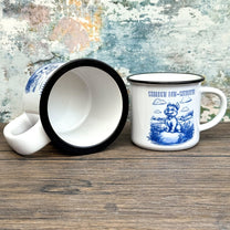 The Quirkify Stardew Valley Chinoiserie Campfire Mug is a blue and white toile ceramic cup featuring house, tree, and mountain artwork with "Stardew Valley" printed on it, displayed on a wooden surface against a multicolored wall.