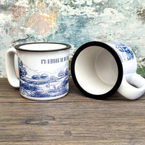 The Quirkify Stardew Valley Chinoiserie Campfire Mug is a blue and white toile ceramic cup featuring house, tree, and mountain artwork with "Stardew Valley" printed on it, displayed on a wooden surface against a multicolored wall.