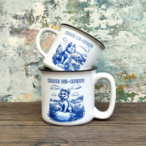 The Quirkify Stardew Valley Chinoiserie Campfire Mug is a blue and white toile ceramic cup featuring house, tree, and mountain artwork with "Stardew Valley" printed on it, displayed on a wooden surface against a multicolored wall.