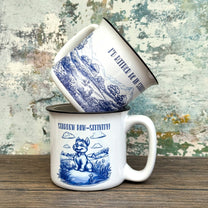 The Quirkify Stardew Valley Chinoiserie Campfire Mug is a blue and white toile ceramic cup featuring house, tree, and mountain artwork with "Stardew Valley" printed on it, displayed on a wooden surface against a multicolored wall.