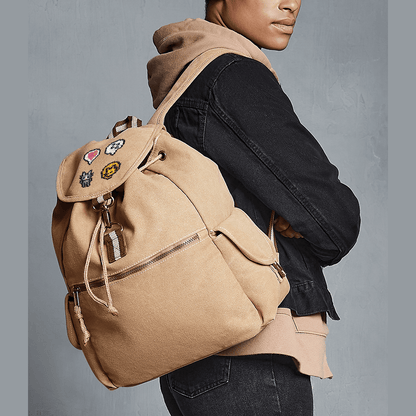 A person wears a tan canvas Stardew Valley backpack adorned with several embroidered patches including a heart emote and cat design.