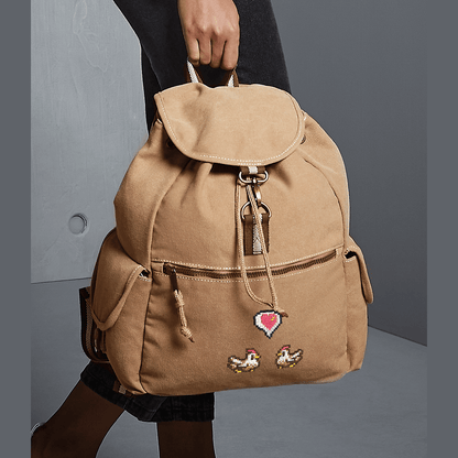 A tan canvas backpack features embroidered chickens and a heart emote referencing the Stardew Valley video game.