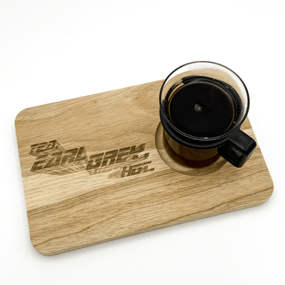A wooden chopping board displays "Tea, Earl Grey, Hot" in a stylized font and holds a glass mug filled with dark liquid.