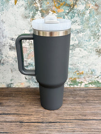 A gray 40oz tumbler with a handle sits on a wooden surface against a textured wall.