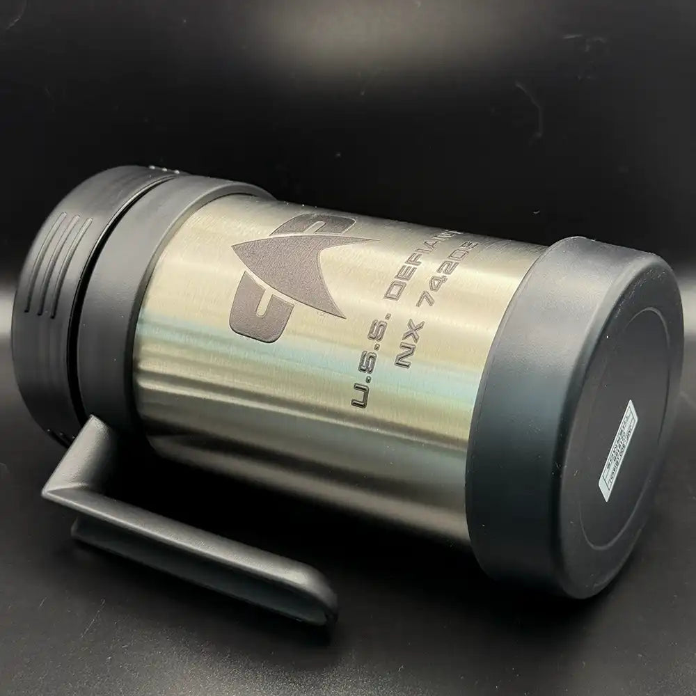 A silver travel mug resembling a thermos features the Star Trek delta shield logo and "U.S.S. Defiant NX 74205" printed on its surface
