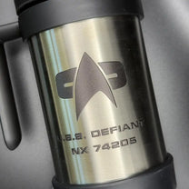 A silver travel mug resembling a thermos features the Star Trek delta shield logo and "U.S.S. Defiant NX 74205" printed on its surface