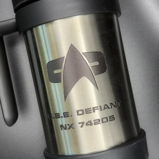 A silver travel mug resembling a thermos features the Star Trek delta shield logo and "U.S.S. Defiant NX 74205" printed on its surface