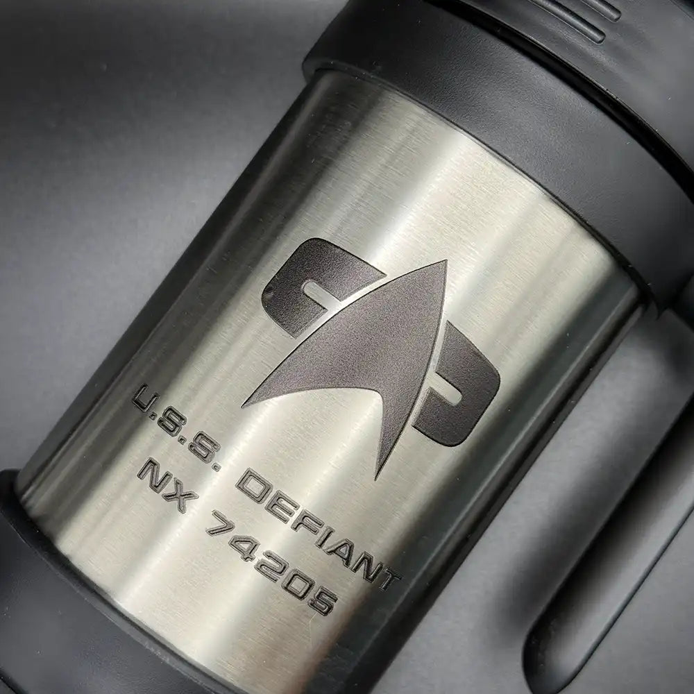 A silver travel mug resembling a thermos features the Star Trek delta shield logo and "U.S.S. Defiant NX 74205" printed on its surface
