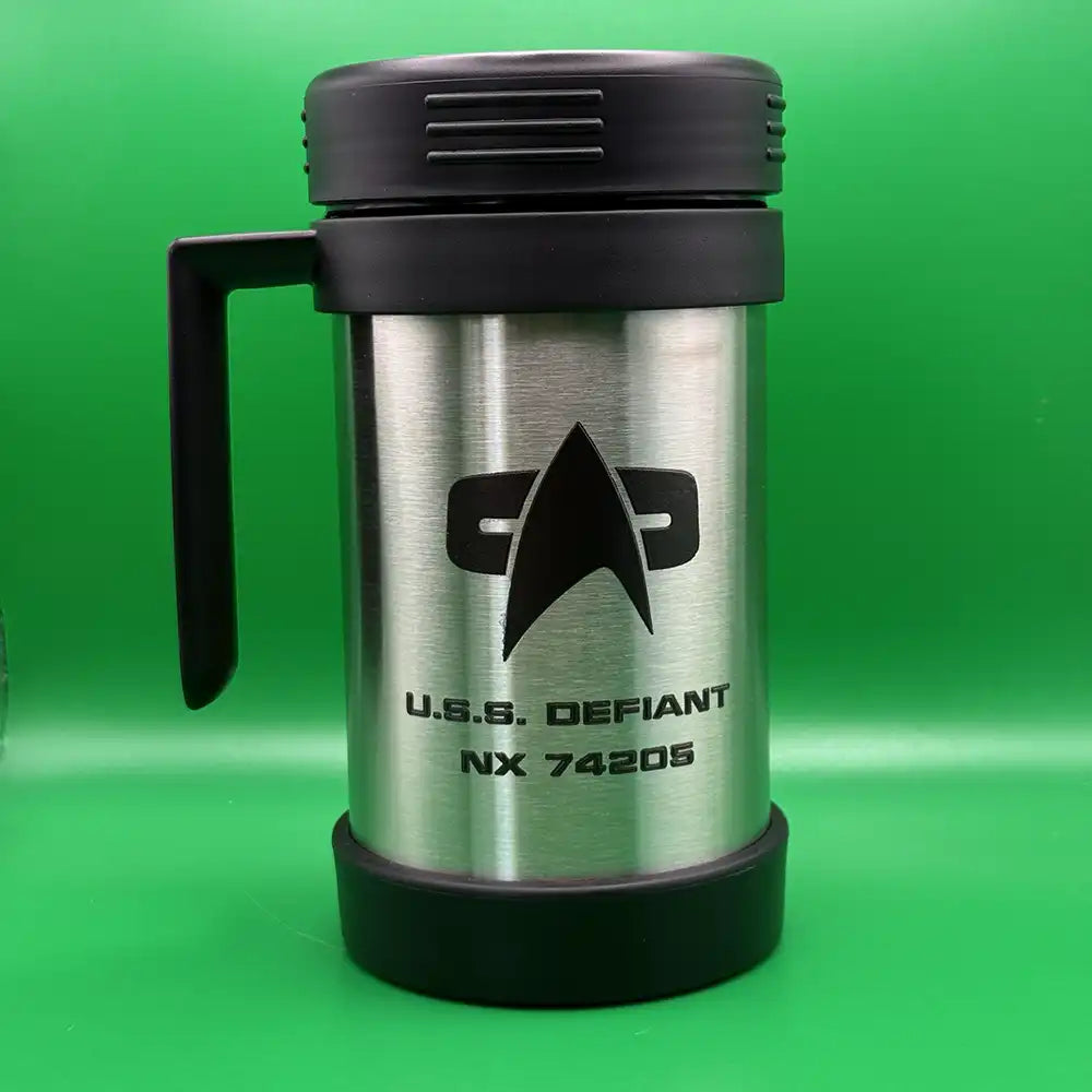 A silver travel mug resembling a thermos features the Star Trek delta shield logo and "U.S.S. Defiant NX 74205" printed on its surface with a green background