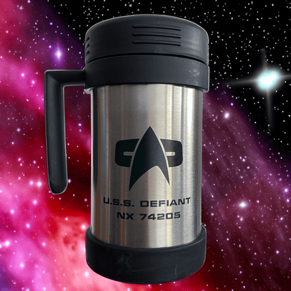 A silver travel mug resembling a thermos features the Star Trek delta shield logo and "U.S.S. Defiant NX 74205" printed on its surface against a backdrop of stars and nebulae.