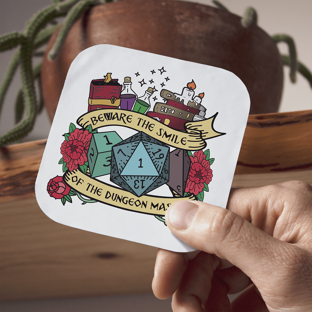 Sticker with dice design and text held by a hand, with a wooden surface and plant in the background