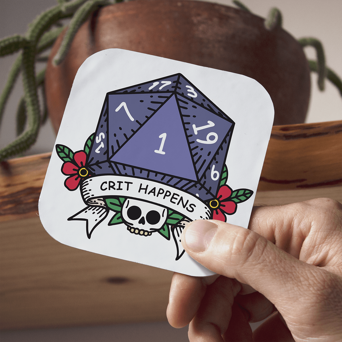 Coaster with a D20 die design, skull, and 'CRIT HAPPENS' text held by a hand.