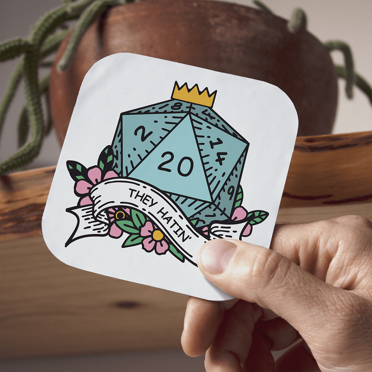 Coaster with a dice design and text held by a hand, with a plant in the background
