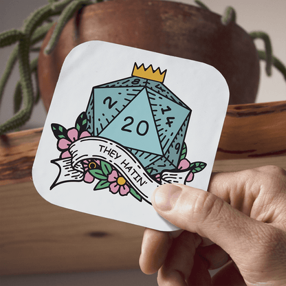 Coaster with a dice design and text held by a hand, with a plant in the background