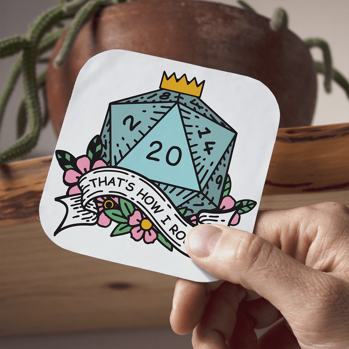Coaster with a DnD D20 die design held by a hand, with a plant in the background