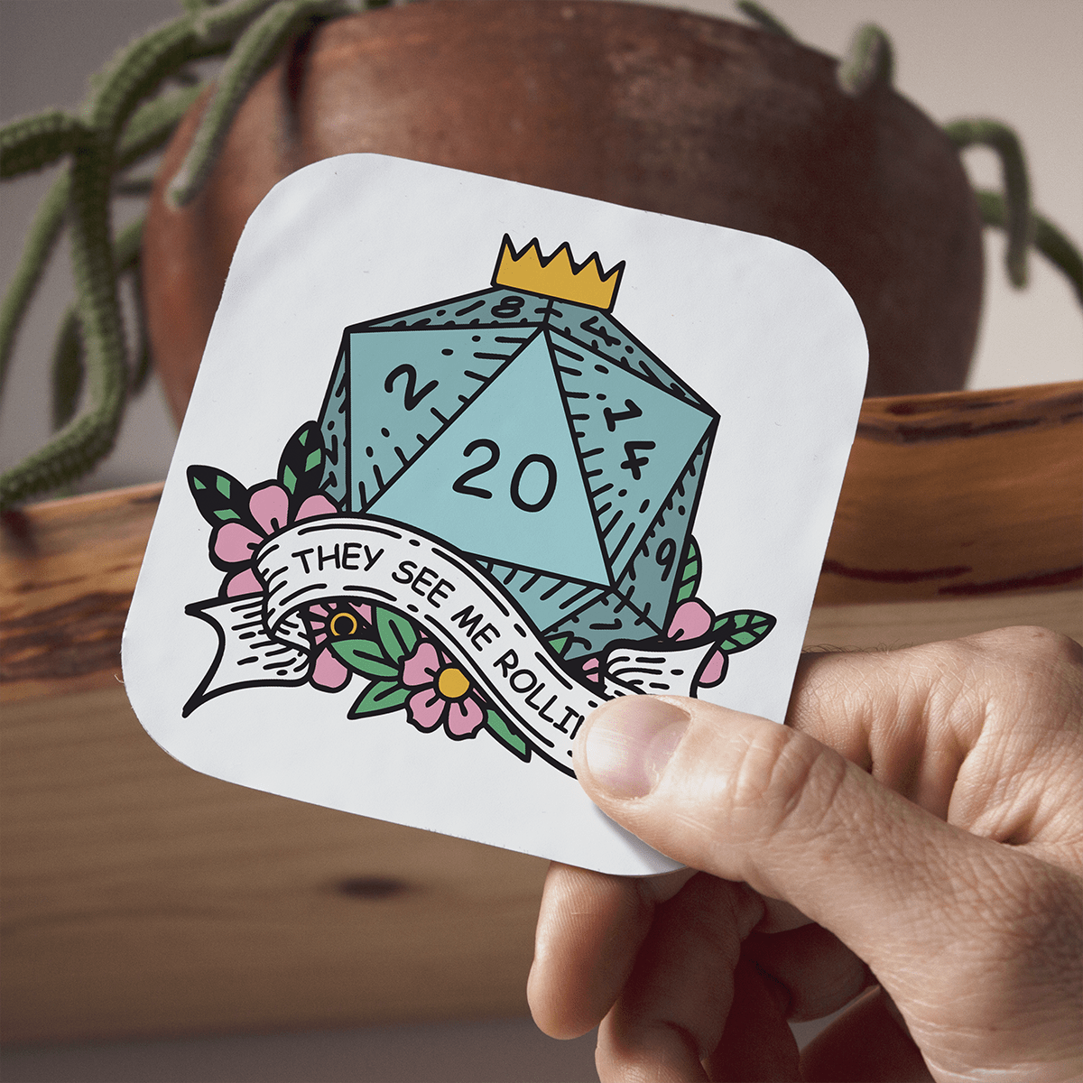 Hand holding a coaster with a D20 DnD die and text, with a plant and pot in the background